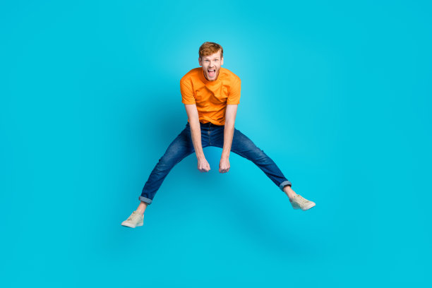 全身照片，印象深刻的红发男孩跳跃，穿着T恤、牛仔裤和运动鞋，背景为蓝色。图片下载