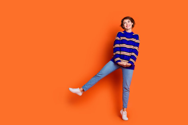 穿着蓝色条纹毛衣和牛仔裤的年轻棕色短发女孩在橙色背景上跳布吉舞的全身照片图片下载