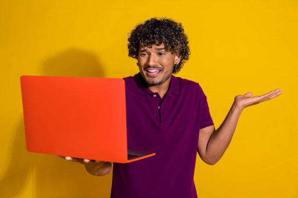 吸引人的年轻男子在黄色背景下穿着时尚的紫色衣服，手持笔记本电脑，表情烦躁地进行视频通话的照片肖像。图片下载