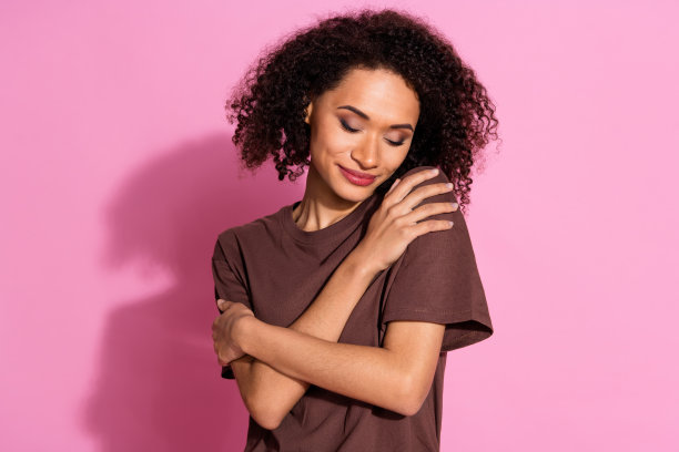 可爱的年轻女士在粉色背景上穿着时尚的棕色服装，闭着眼睛自我拥抱的照片肖像图片下载