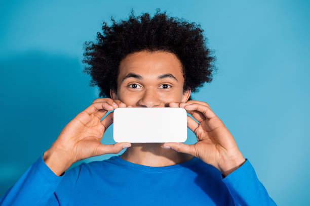 吸引人的年轻男子用手遮住嘴，穿着时尚的衣服，站在蓝色背景前，手持白色屏幕设备的照片肖像。图片下载