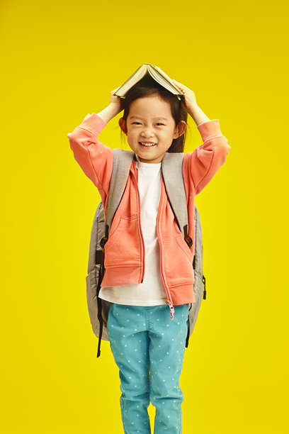 可爱的微笑亚洲女孩，背着书包，手里托着一本打开的书，站在黄色的背景前。图片下载