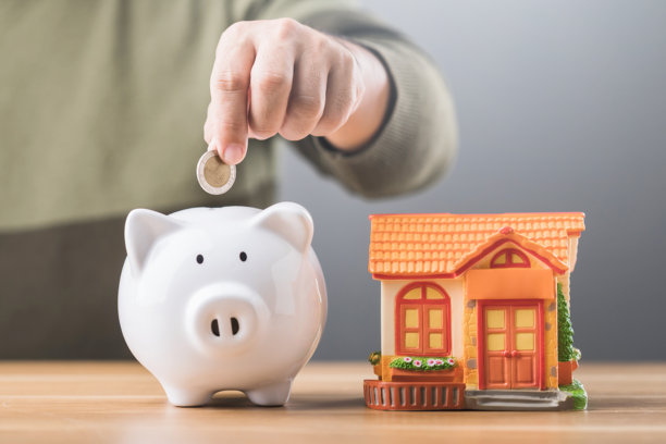 一个男人将硬币投进桌子上的白色储蓄罐，储蓄罐上有房屋模型，象征着为购房或房地产投资而储蓄。这张图片反映了深思熟虑的财务规划。图片下载