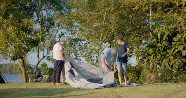 家庭露营者与祖父、母亲和儿子在自然保护区搭帐篷的 candid 照片 - 周末团队合作，在黄昏时分一起享受自然活动的亲密时光。图片下载