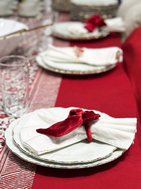 圣诞餐桌摆设的图片，叠放的波浪边餐盘和茶盘，白色餐巾上放着礼物和红色天鹅绒蝴蝶结餐巾环，金色长柄勺子放在碗里，饮用玻璃杯，红色桌布，桌旗。图片下载