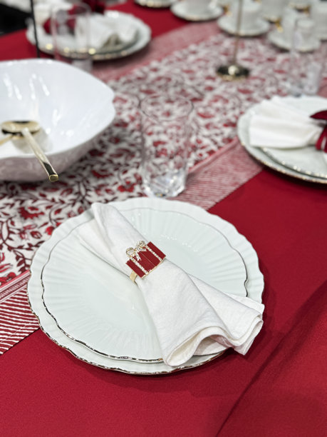 圣诞节餐桌摆设的图片，叠放的波浪边餐盘和茶盘，带有礼物设计的白色餐巾环，金色长柄勺子放在碗里，饮用杯，酒杯，红色桌布配桌旗图片下载