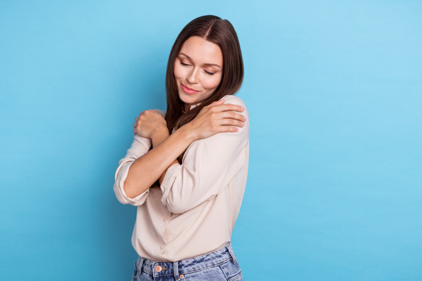 可爱的年轻女士在蓝色背景下拥抱自己的肩膀，展现自我关怀，穿着时尚休闲服装的照片肖像。图片下载
