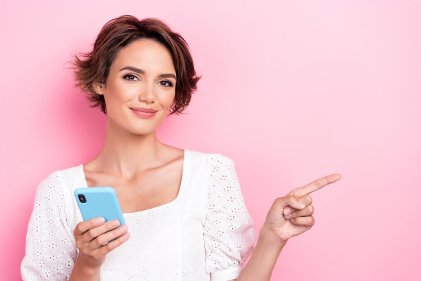 年轻女士的照片肖像，手持金色智能手机，展现出时尚感，穿着白色服装，背景为粉色，留有空白空间。图片下载