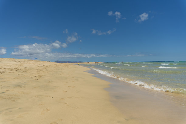 令人惊叹的西班牙加那利群岛富埃特文图拉岛索塔文托海滩的美景。照片捕捉了广阔的碧蓝海水、金色沙丘以及远处戏剧性的火山景观。图片下载