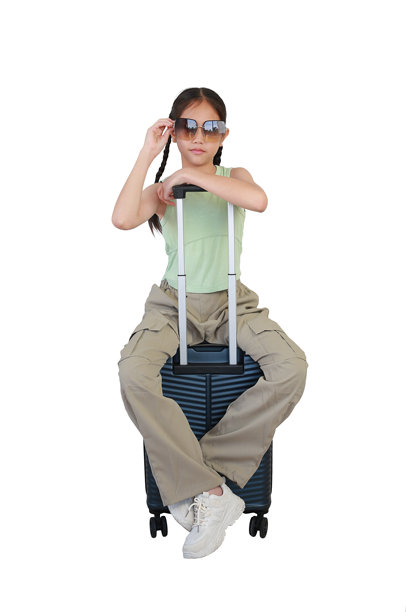 亚洲女孩坐在行李箱上，摆姿势并直视镜头，佩戴着太阳镜，背景为白色。儿童时尚与旅行概念。全身照片。图片下载