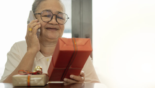 一位安详的亚洲老年女士坐着，使用智能手机，身边有圣诞礼物的头像照片。图片下载