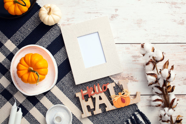 秋季平铺布局，包含照片框模型、南瓜、棉花枝、蜡烛和“你好，秋天”标志，放置在木桌上的格子布上。俯视图图片下载