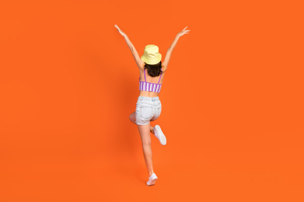 穿着紫色泳衣的幽默无忧女士在橙色背景下举起双臂的全身照片图片下载