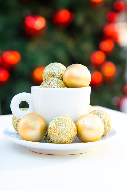 咖啡杯内外装饰着金色圣诞球的竖版照片。白色桌子上的咖啡搭配，灰色椅子，背景是挂有红色玩具的新年树。冬季咖啡馆。明信片风格的图片。图片下载