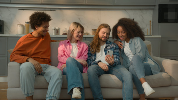 白人男性手持手机，向多种族朋友展示搞笑图片或幽默视频，大家在沙发上欢笑，享受快乐时光。图片下载