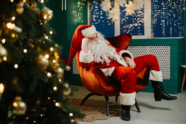 疲惫的圣诞老人坐在装饰华丽的圣诞树旁的扶手椅上休息，经过了一整天的送礼。图片下载