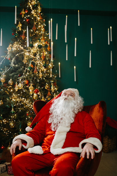 疲惫的圣诞老人坐在装饰华丽的圣诞树旁的扶手椅上休息，经过了一整天的送礼。图片下载