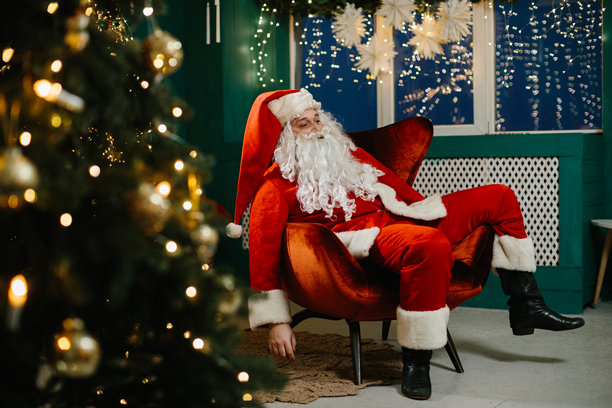 疲惫的圣诞老人坐在装饰华丽的圣诞树旁的扶手椅上休息，经过了一整天的送礼。图片下载