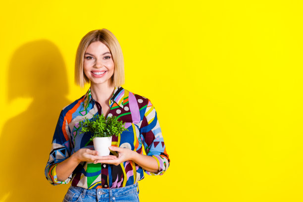 时尚多彩服装的年轻女性手持室内植物的照片肖像，背景为黄色。图片下载