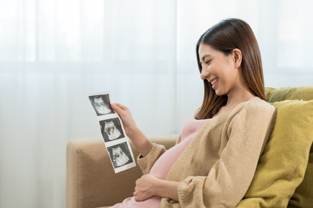 快乐的年轻孕妇在家中看着超声波扫描照片，轻抚着自己的肚子，期待着幸福的母亲时光，准备迎接孩子的出生，进行家庭规划并为孩子的健康投保。图片下载