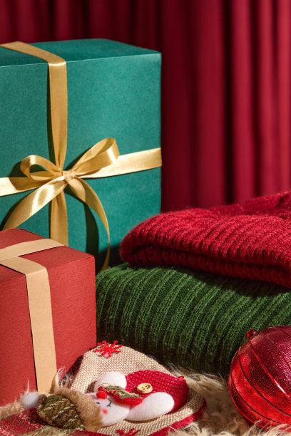 折叠的毛衣放在毛皮地毯上，旁边是礼品盒、雪人袜子和圣诞球。背景中的红色窗帘为照片营造了温暖的圣诞氛围。图片下载