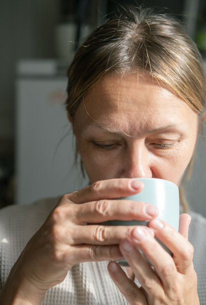 的白人女性在家中窗边喝茶或咖啡。家庭生活方式的概念。阳光明媚的日子里，穿着浴袍的现代快乐中年女士的特写照片。图片下载