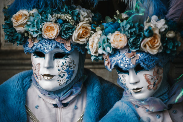一对情侣的头像肖像，身穿协调的白色和蓝色服装，头上戴着花朵，背景是威尼斯狂欢节。图片下载
