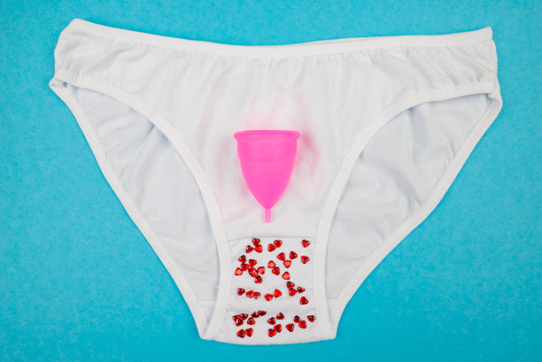 白色棉质经典内裤的俯视照片，上面有红色心形纸屑和月经杯，背景为独特的浅蓝色。图片下载