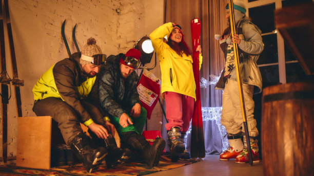 团队在室内准备滑雪，系紧靴子并检查装备，穿着鲜艳的服装，周围布置着节日的灯光和装饰。图片下载