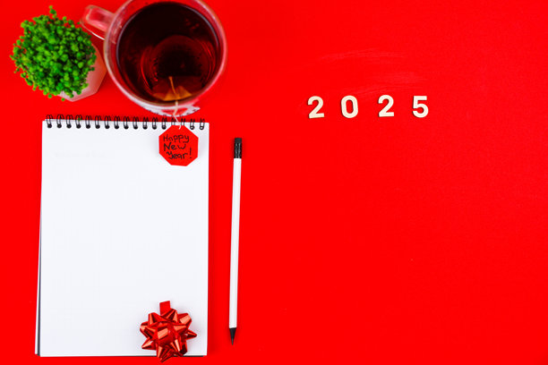 一只白色的笔记本上有一个黑色的弹簧，上面放着一个鲜红色的礼品蝴蝶结，照片的右侧还有一支白色的铅笔，旁边是一盆绿色植物和一杯带有红色“新年快乐”标签的茶，以及木制数字。图片下载