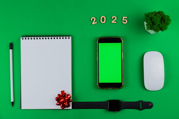 照片中央是一部黑色手机，屏幕为绿色抠像，周围有一本黑色螺旋装订的白色笔记本、一支带黑色橡皮和铅芯的白色铅笔、木制数字2025、一盆绿色办公植物、一只白色电脑鼠标和一部黑色设备。图片下载