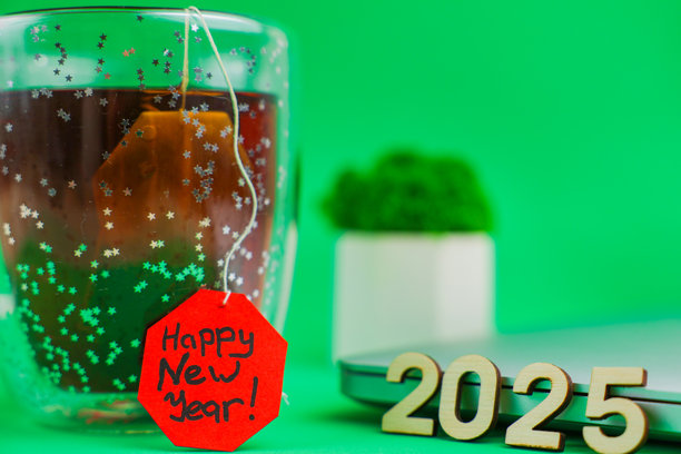 照片前景侧面有一个透明的闪光杯，里面装着黑茶茶包，茶包的标签是鲜红色，上面写着“新年快乐”。旁边放着一台关闭的笔记本电脑，电脑上摆放着木质数字2025。图片下载