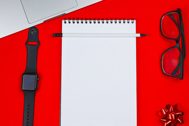 一只白色笔记本配有黑色弹簧，旁边是一支黑色铅芯和橡皮的白色铅笔，置于红色背景的照片中央，周围有一块黑色手腕智能手表、一台笔记本电脑、黑框眼镜和一个节日红色蝴蝶结。图片下载