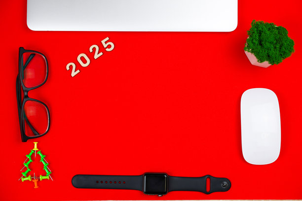一台灰色的关闭笔记本电脑，旁边有木制数字“2025”，位于红色背景的照片上，旁边有一棵由绿色文具图钉组成的创意圣诞树，背景同样是红色，还有一副黑框眼镜。图片下载