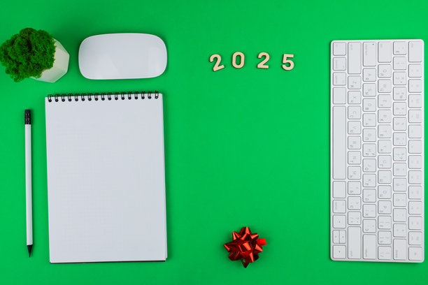 一台白色的英式电脑键盘飞向照片的右侧，上面放着木制数字2025、一盆绿色办公植物和一只白色电脑鼠标，左侧是一只带黑色弹簧的白色笔记本和一支黑色铅芯的白色铅笔。图片下载