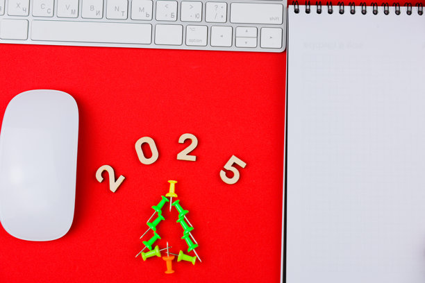 在红色背景的照片中央，有木制数字2025和用办公钉制作的圣诞树，旁边有一本顶部带黑色弹簧的白色笔记本和一个电脑鼠标，上面放着一个白色键盘。图片下载