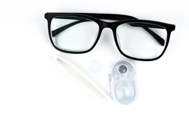 照片中央是黑框眼镜的特写，眼镜后面是一次性隐形眼镜容器，蓝色镜片和米色镊子的特写，背景为白色，留有文字空间。视力、矫正和眼科。图片下载