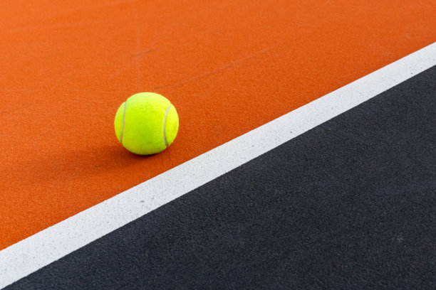 一张垂直照片，展示了一颗黄色网球在橙色和黑色的网球场上 - 乒乓球场，白色的底线和黑色的界外区域。图片下载
