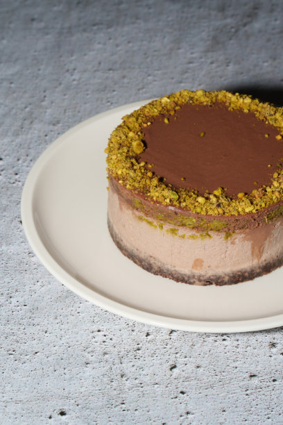 迪拜巧克力奶酪蛋糕，夹心开心果酱和卡塔伊菲，外层覆盖巧克力甘纳许，摆放在白色盘子上。灰色背景留有空白。竖版照片。奢华，精致甜点。图片下载