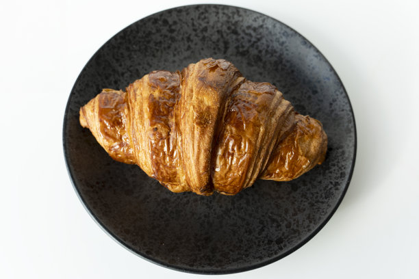 新鲜出炉的可颂面包放在黑色盘子上。单个法式糕点特写照片。涂了黄油的可颂面包放在黑色盘子上。图片下载