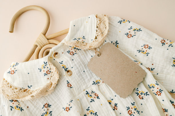 特写镜头展示了一件带有蕾丝细节的花卉婴儿裙，配有环保的牛皮纸标签，背景为柔和的米色。这张图片强调了可持续性和复古风格的婴儿服装设计。图片下载