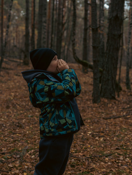 一个迷路的孩子穿着温暖的冬季外套和帽子，用手捂住嘴巴在黑暗的秋季森林中呼喊求助。一个害怕的孩子在森林里迷路了，正在呼喊求助。图片下载