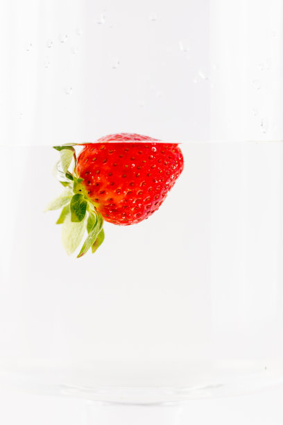 在白色背景的照片上，一颗成熟的草莓浸泡在清水中，洗净了浆果和水果，富含健康维生素的季节性夏季水果，草莓的绿色果梗像一顶皇冠。图片下载
