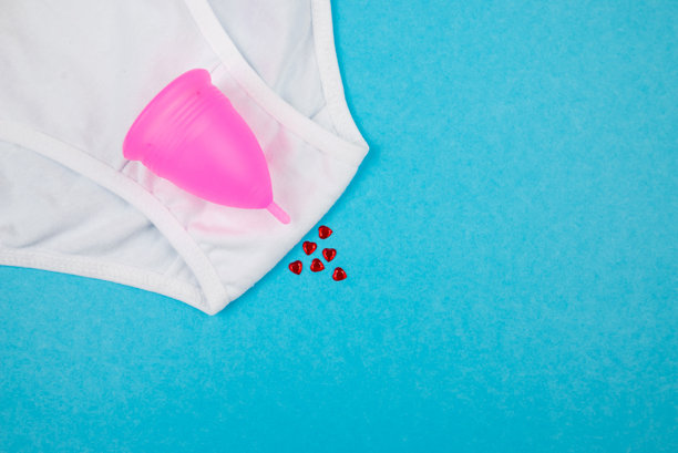 白色棉质经典内裤的俯视照片，上面有红色心形纸屑和月经杯，背景为独立的柔和蓝色。图片下载