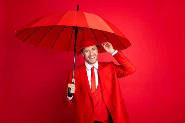 帅气年轻男士在红色背景下触碰帽子，手持雨伞，穿着时尚经典的服装，拍摄的肖像照片。图片下载