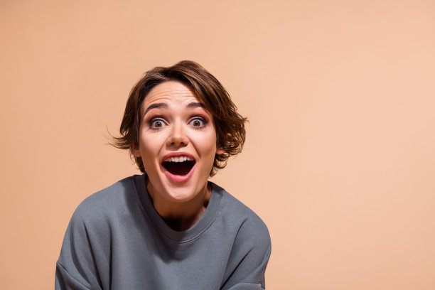 漂亮年轻女孩的照片肖像，惊讶的表情，张嘴，穿着时尚的灰色服装，背景为米色。图片下载
