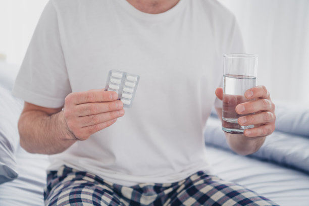 年轻男子穿着睡衣，手持水杯和药片，醒来后在现代舒适的室内卧室中拍摄的特写照片图片下载