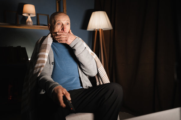 一个孤独的退休老人晚上独自坐在家里，看电视，看到的内容让他感到震惊，手捂住了嘴。图片下载