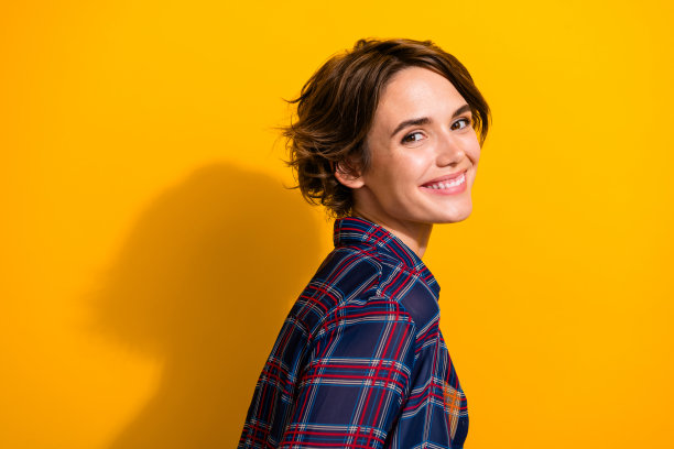 侧面照片，露齿而笑的女孩，短发，穿着格子衬衫，面向镜头，背景为黄色。图片下载