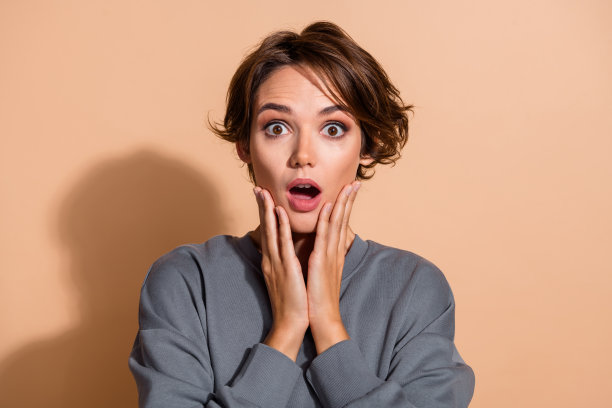 漂亮年轻女孩的照片肖像，惊恐的表情，手触脸，穿着时尚的灰色服装，背景为米色。图片下载